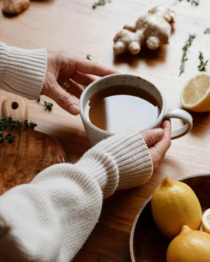 Person holding a speckled mug of warm broth on wooden table with fresh ginger, lemon, and herbs nearby, cozy setting with Beef Bone Broth Concentrate - Natural - 250g