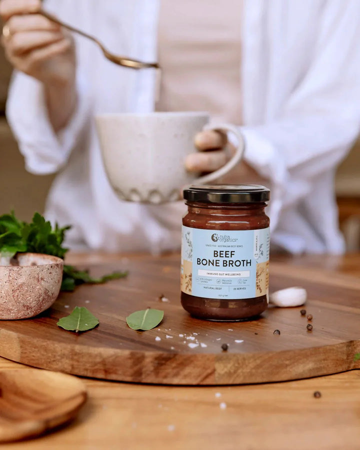 Beef Bone Broth Concentrate - Natural - 250g jar on wooden board with herbs and person holding cup in background