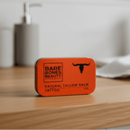 Tattoo Tallow Balm - 40ml by Bare Bones Beauty in an orange metal tin on a wooden surface with blurred background.